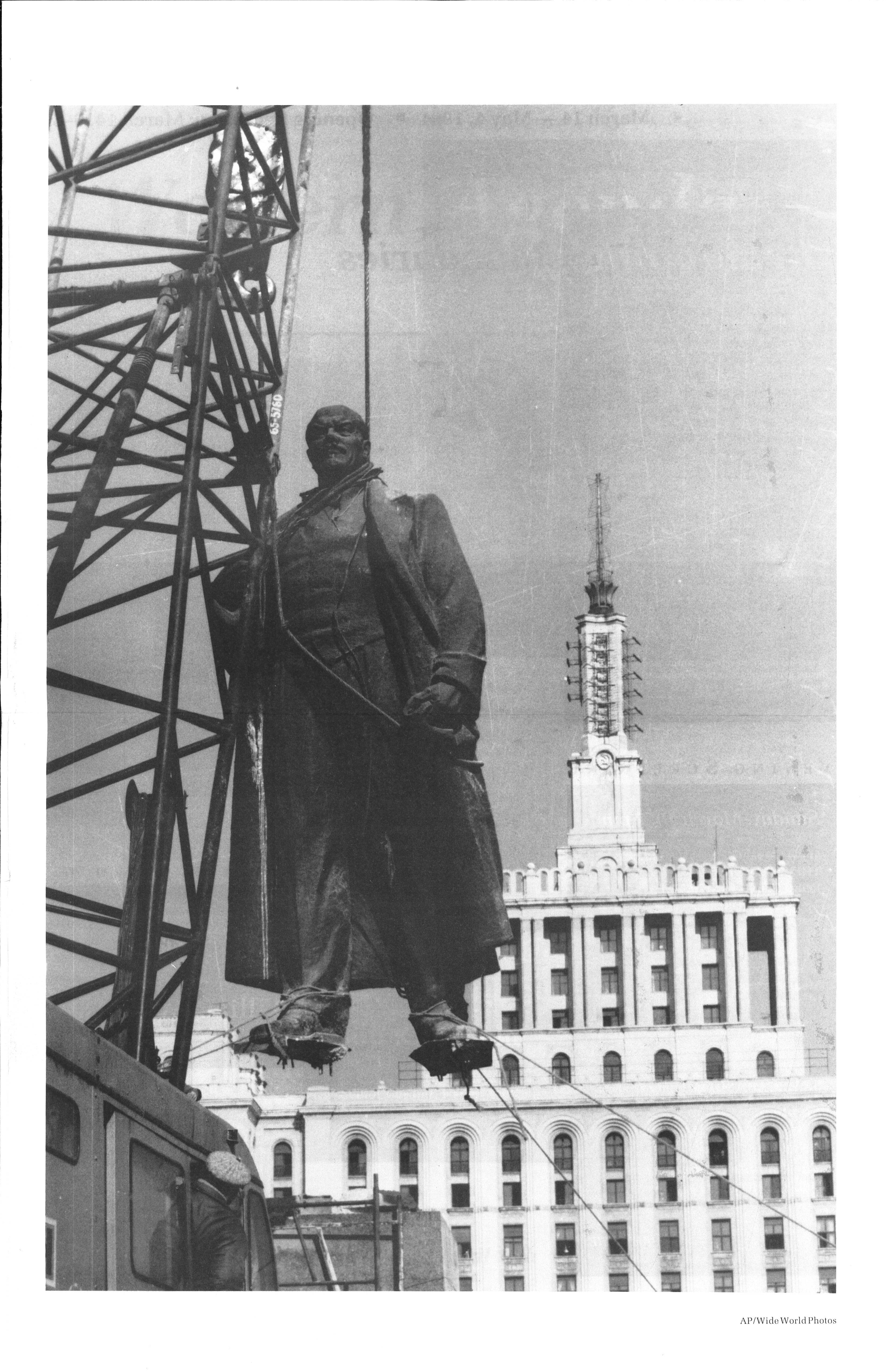 A metal statue is being lifted from its platform by a crane via ropes around its neck and feet. A large building is visible behind it.