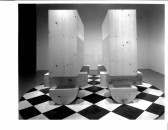 Two identical large wooden sculptures on a black and white checkered floor. The sculptures have rounded, semi-circle shaped bases with rectangular pieces set crosswise atop that.