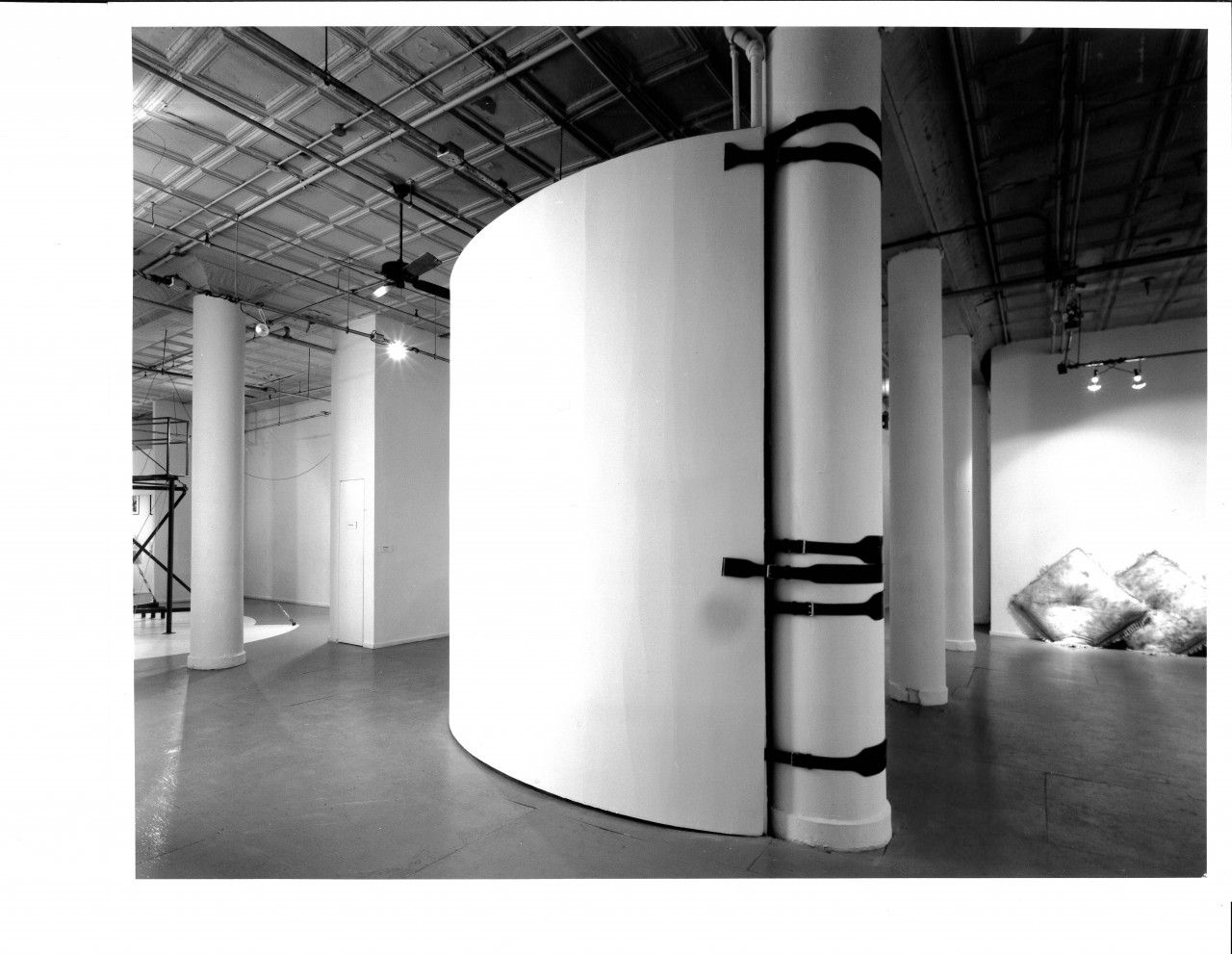 A black and white photograph of a large convex panel strapped to a white column, its circular side extending out into an empty room.