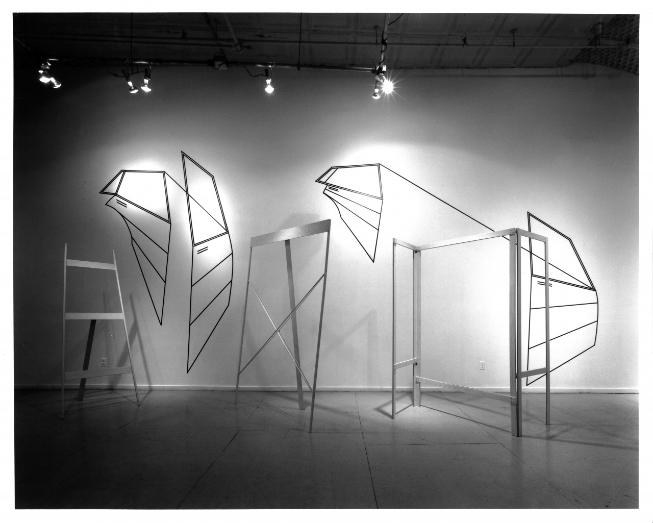 A black and white photograph of a sculpture lit from above. It has several suspended shapes resembling detached car doors, with additional minimal pieces.