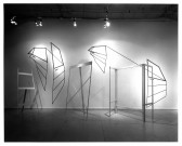 A black and white photograph of a sculpture lit from above. It has several suspended shapes resembling detached car doors, with additional minimal pieces.