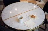 A round, diner-style table with a white mug, napkins, a baked good sitting on top of its open wrapper, and a small card with text. A long, straight line of crumbs stretches across the diameter of the table.