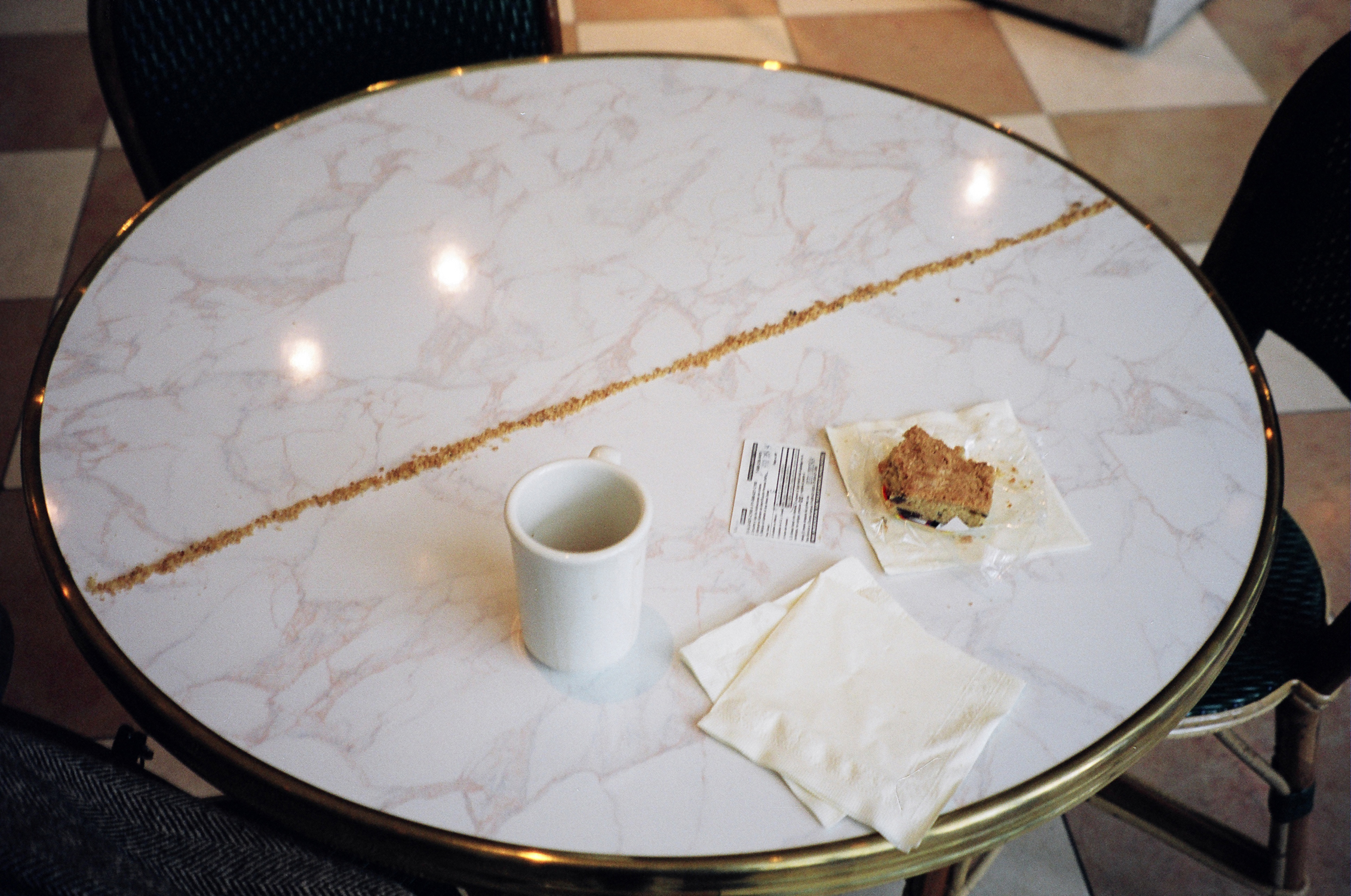 A round, diner-style table with a white mug, napkins, a baked good sitting on top of its open wrapper, and a small card with text. A long, straight line of crumbs stretches across the diameter of the table.