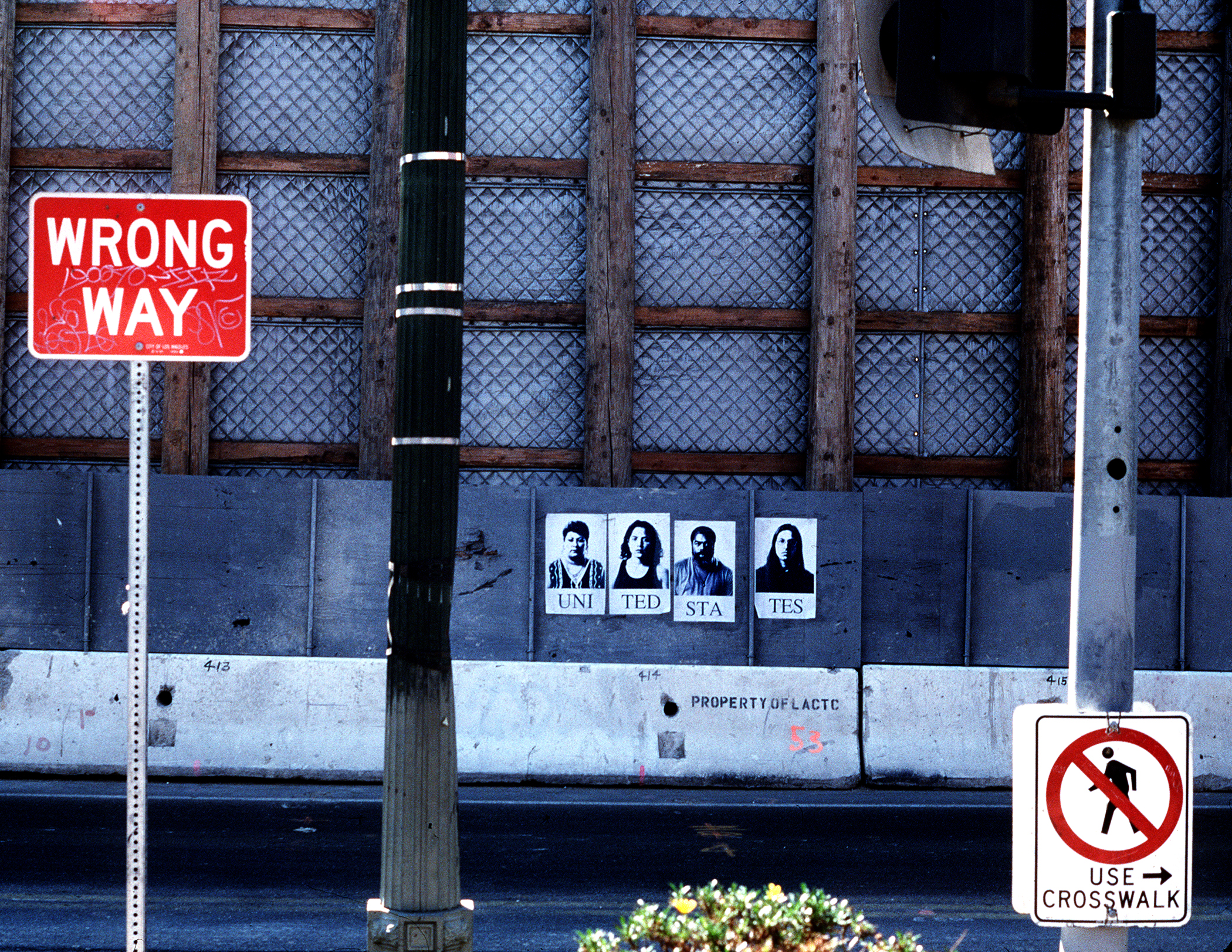 A city street with a concrete median and wall made from gray metal supported by wooden planks. The median has the words 