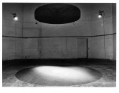A black and white image of a large room with a round black disk suspended from the ceiling over a gray circle in the floor. It is lit from above by mounted lights.