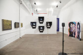 Several artworks installed in a gallery space with white walls. On a wall to the left of the image, three yellow toned paintings are installed at eye level. On a wall at the far center of the image, four black viewing boxes are installed on the wall in a diamond formation. On the gallery wall to the right, two painted portraits sit on the ground.