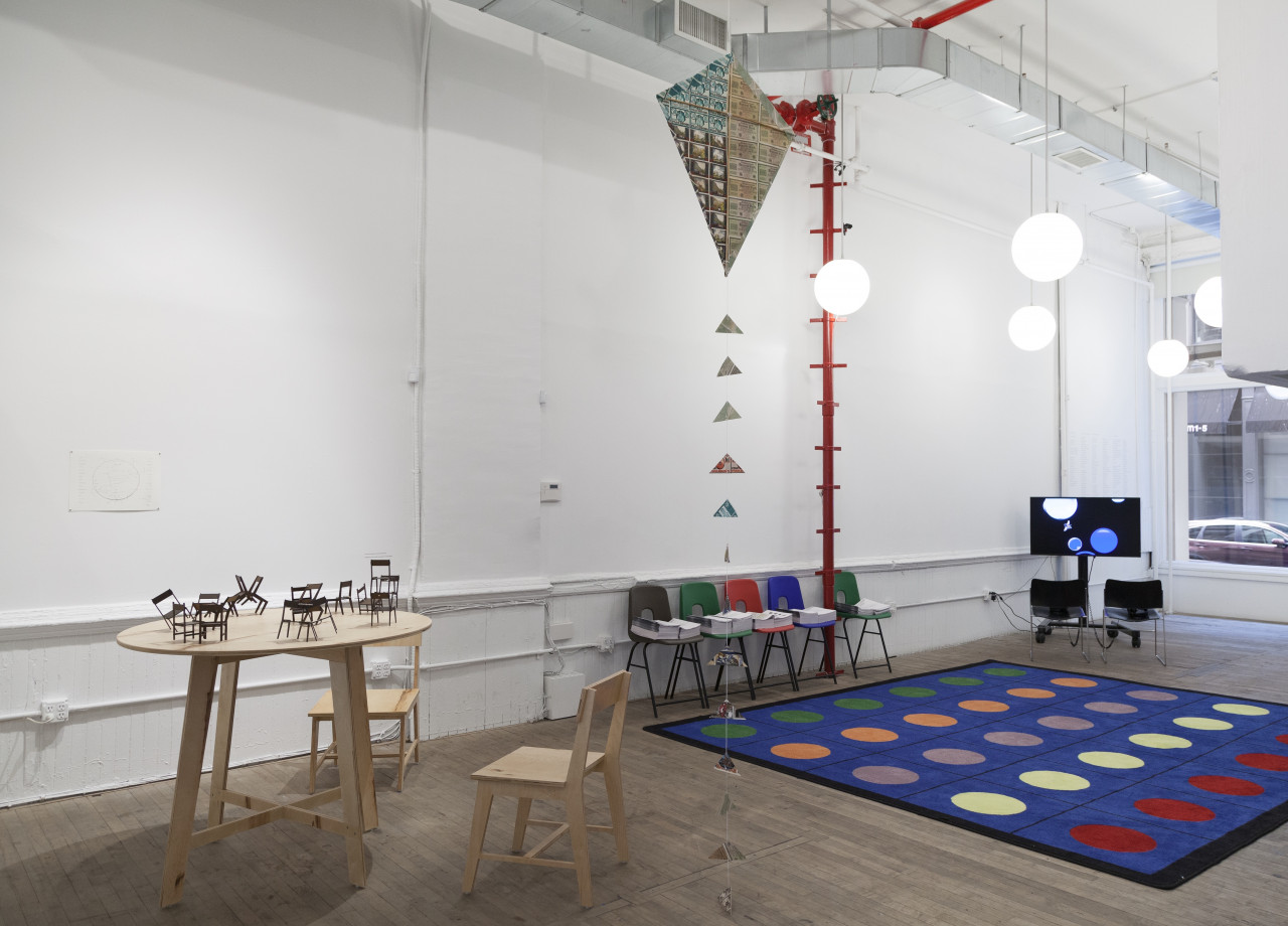 A wooden table with two chairs arranged in a gallery space. A kite hangs from the ceiling to the right of the chair and table set. Behind the kite is a large, blue carpet spread across the floor, several colorful plastic chairs lined up against the wall, and a video monitor.