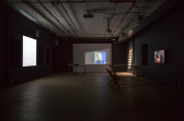 Three projected images illuminate three disparate walls of a darkened room. The image on the left wall is white, while the image on the far center wall shows a figure who appears to be singing against a blue and green background. The image on the right wall shows two figures wearing headphones with their backs turned from the viewer.