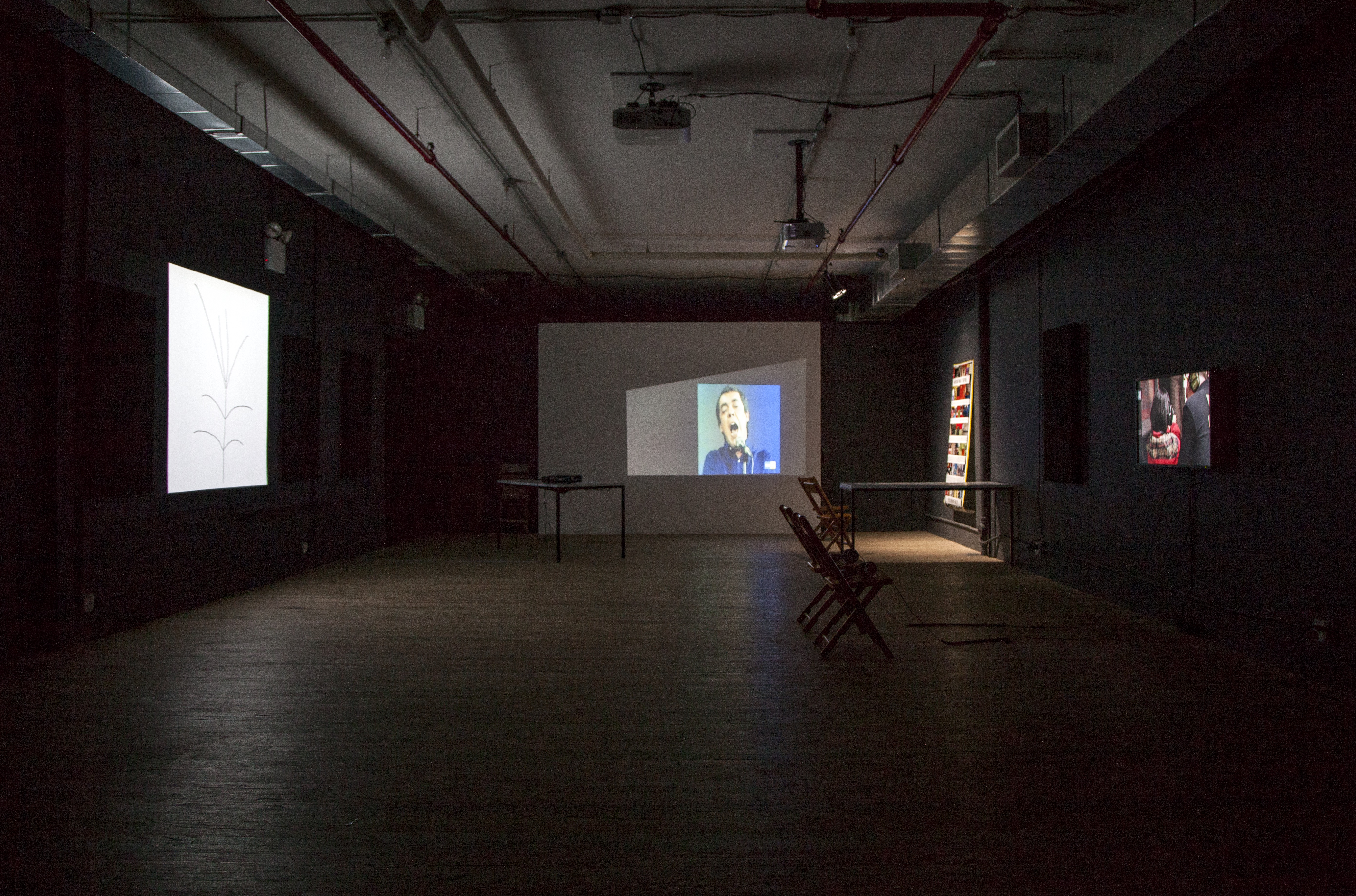 Three projected images illuminate three disparate walls of a darkened room. The image on the left wall is white, while the image on the far center wall shows a figure who appears to be singing against a blue and green background. The image on the right wall shows two figures wearing headphones with their backs turned from the viewer.