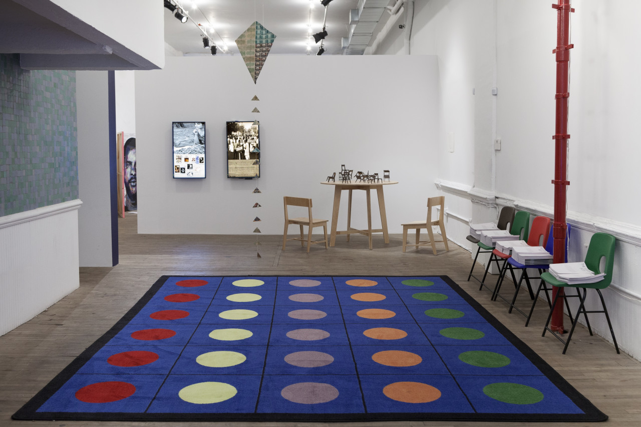 A blue carpet decorated with colorful polka-dots fills the floor of a gallery space. Colorful, plastic chairs are arranged against the right wall. A wooden table with chairs and two monitors are displayed at the space
