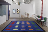 A blue carpet decorated with colorful polka-dots fills the floor of a gallery space. Colorful, plastic chairs are arranged against the right wall. A wooden table with chairs and two monitors are displayed at the space's back wall. A kite hangs from the ceiling in the center of the space.
