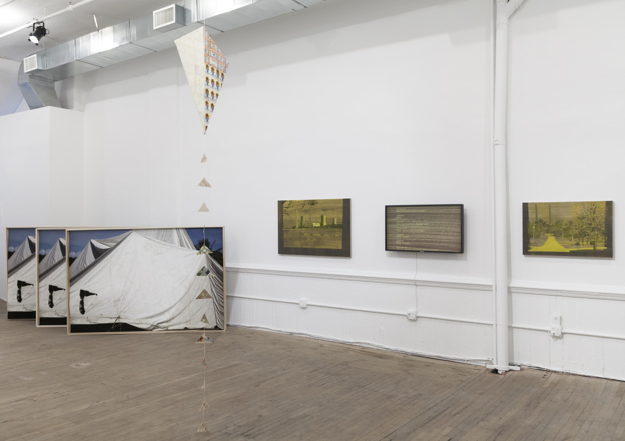 A kite hangs from the ceiling of an exhibition space in front of a white gallery wall. Three yellow-toned paintings are displayed at eye level on the wall behind the kite. Three identical paintings depicting an image of a white, tent-like tarp sit at an angle on the gallery floor to the left of the yellow paintings.