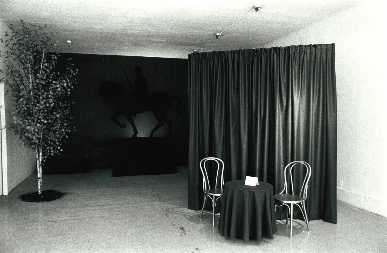 A round table and two chairs sit in front of a tall curtain. To the right, a tree extends from the floor to the ceiling. In the background of the image is a sculpture of a man on a horse.
