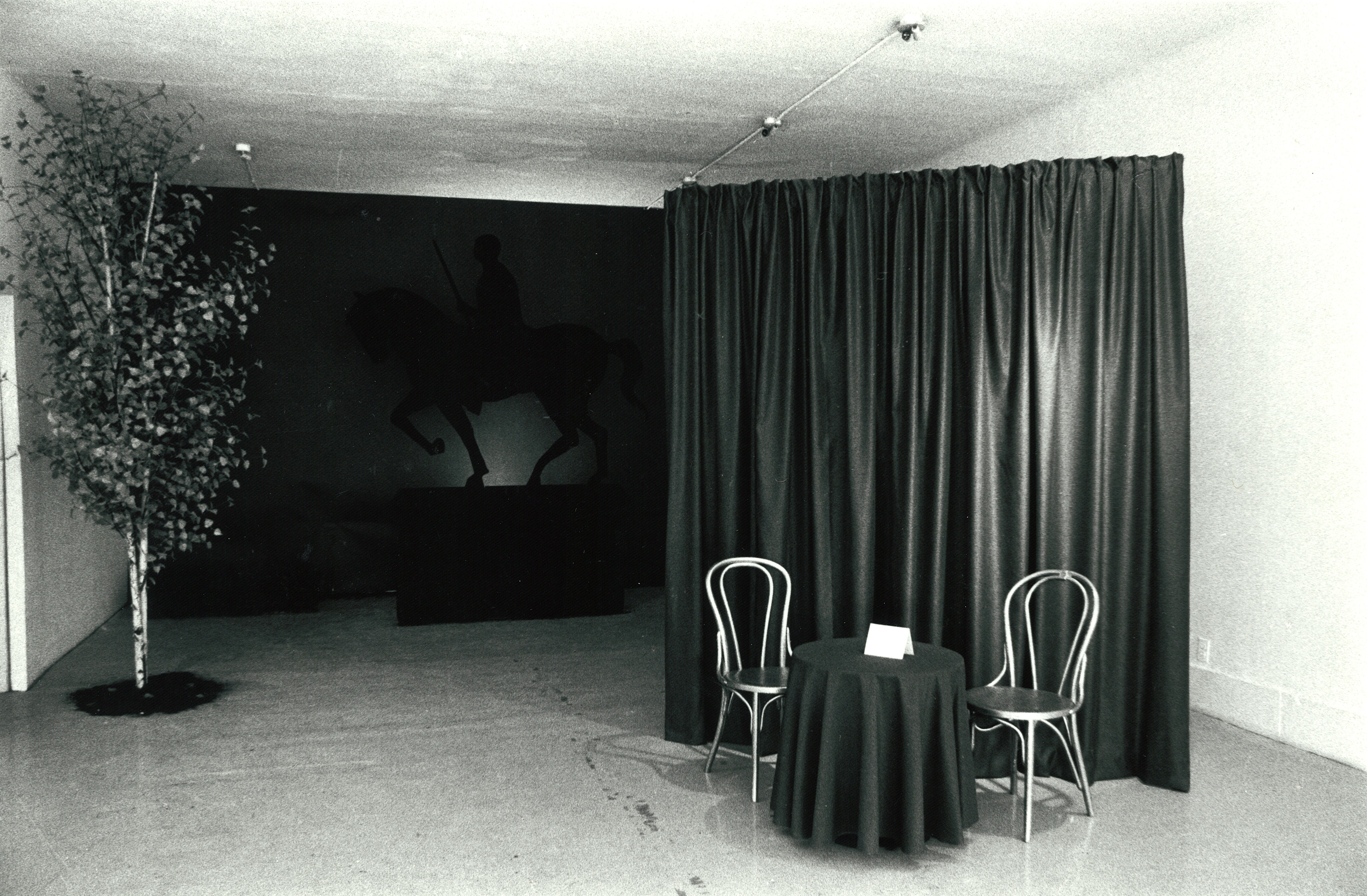 A round table and two chairs sit in front of a tall curtain. To the right, a tree extends from the floor to the ceiling. In the background of the image is a sculpture of a man on a horse.