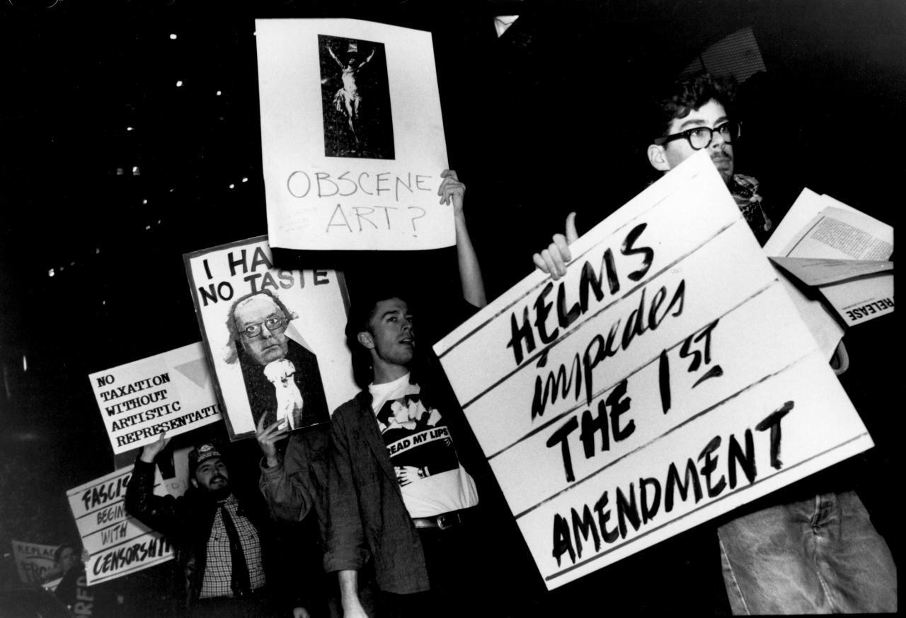 A line of people hold signs with messages including, "ED HELMS impedes THE 1ST AMENDMENT" and "OBSCENE ART?".