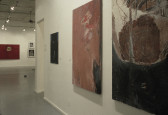 A close up view of two abstract, earth-toned paintings hanging on a wall.  Beyond them a doorway and another room with more painting is visible.