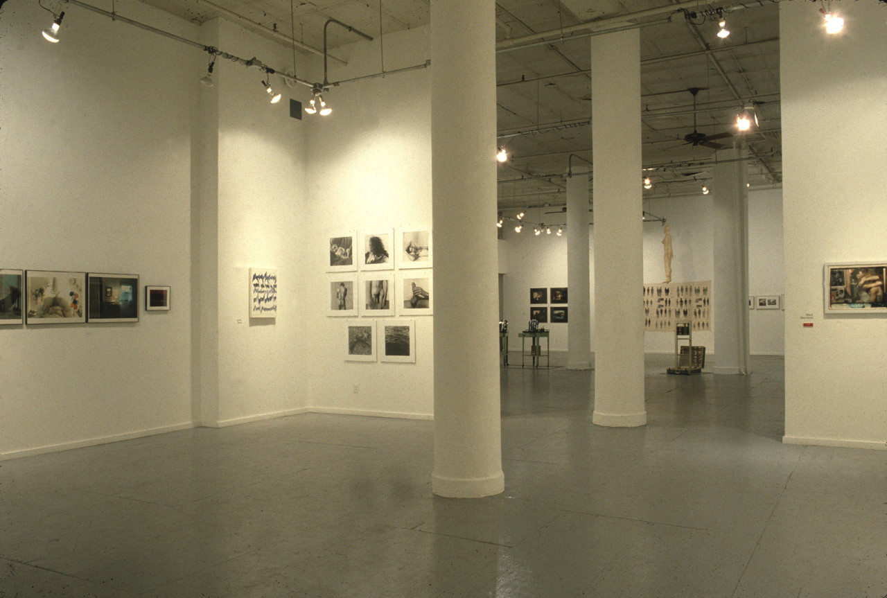 Two rooms in a large gallery space are visible, with artworks hung on the walls and sculptures in the center of the further room.