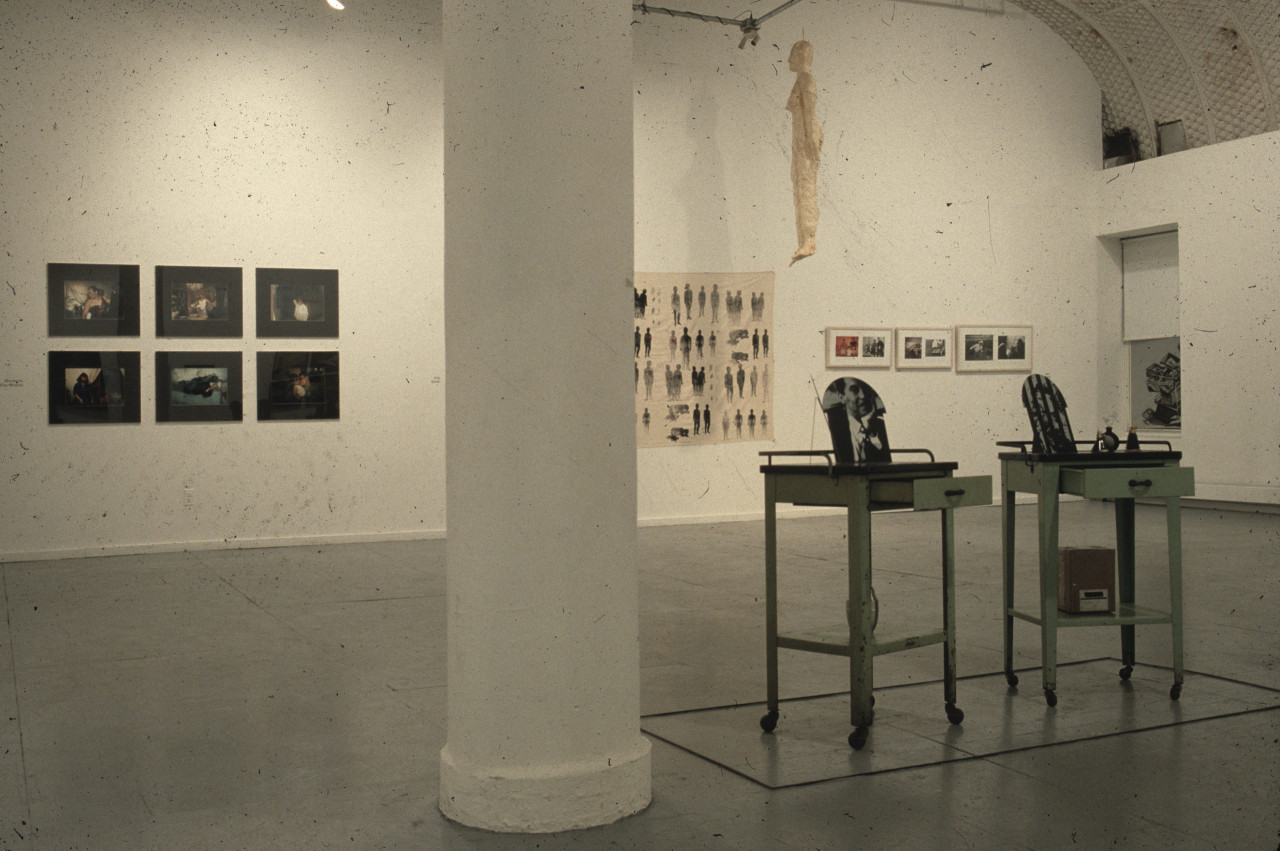 A wide view of a gallery.  A sculpture including two small tables is next to a column in the center of the room.  The far wall is hung with paintings and photographs, and a monochormatic white sculpture of a human figure hangs from the ceiling.