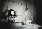 Black-and-white photograph of a TV against a brick wall, connected to a camera recording directly above a spotlighted chair and desk.