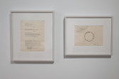Color photograph of two framed works that are hung on the wall. The piece on the left contains typed instructions for a musical performance, and the piece on the left contains hand-written notes for another performance.
