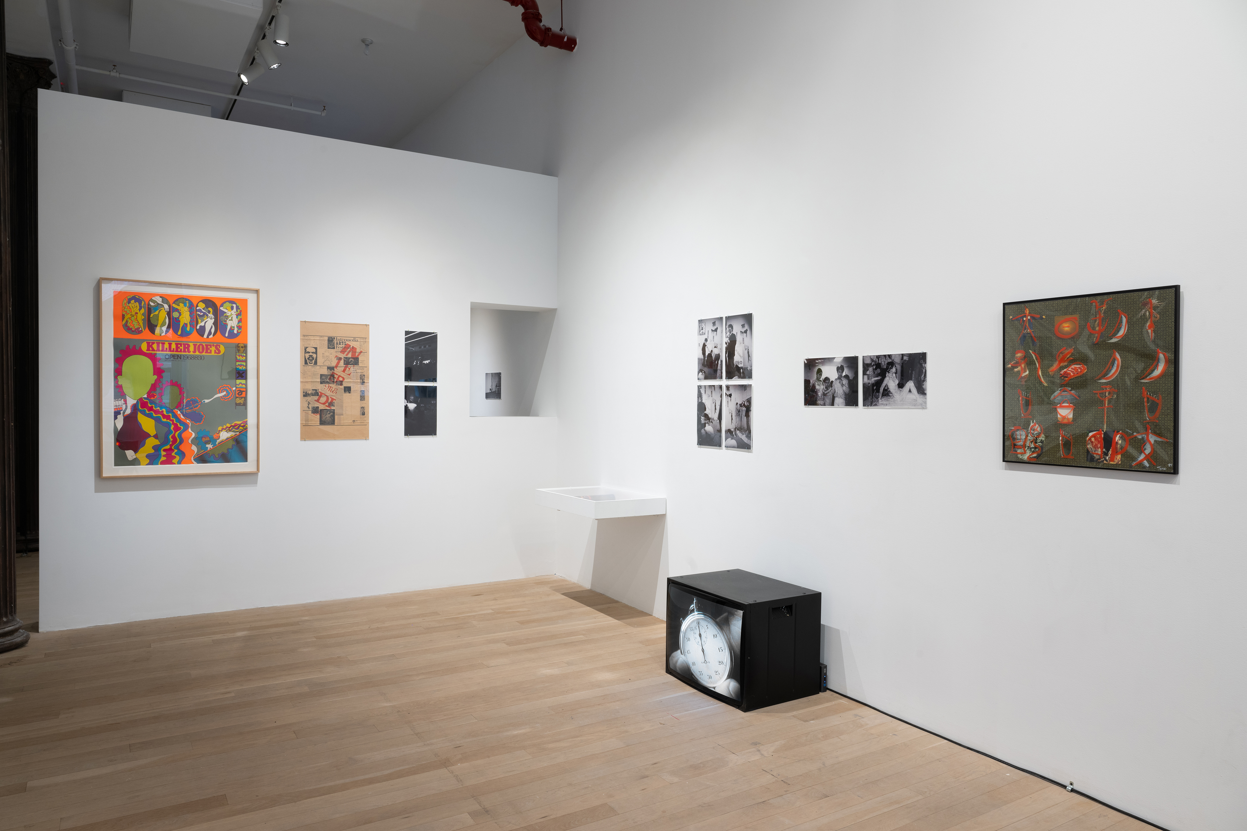 Color photograph of a corner of a gallery space. On the left wall hang various framed posters, and black-and-white photographs. A small window in the wall leads to the front of the gallery. On the right wall are more photographs and posters, and a vitrine. On the floor sits a television monitor displaying a black-and-white image of a pocketwatch.