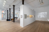Color photograph of a gallery space. Various musical instruments and audio devices are placed around the back room, photographs and vitrines hang on the walls, and various television monitors and projections are placed along the sides of the room.