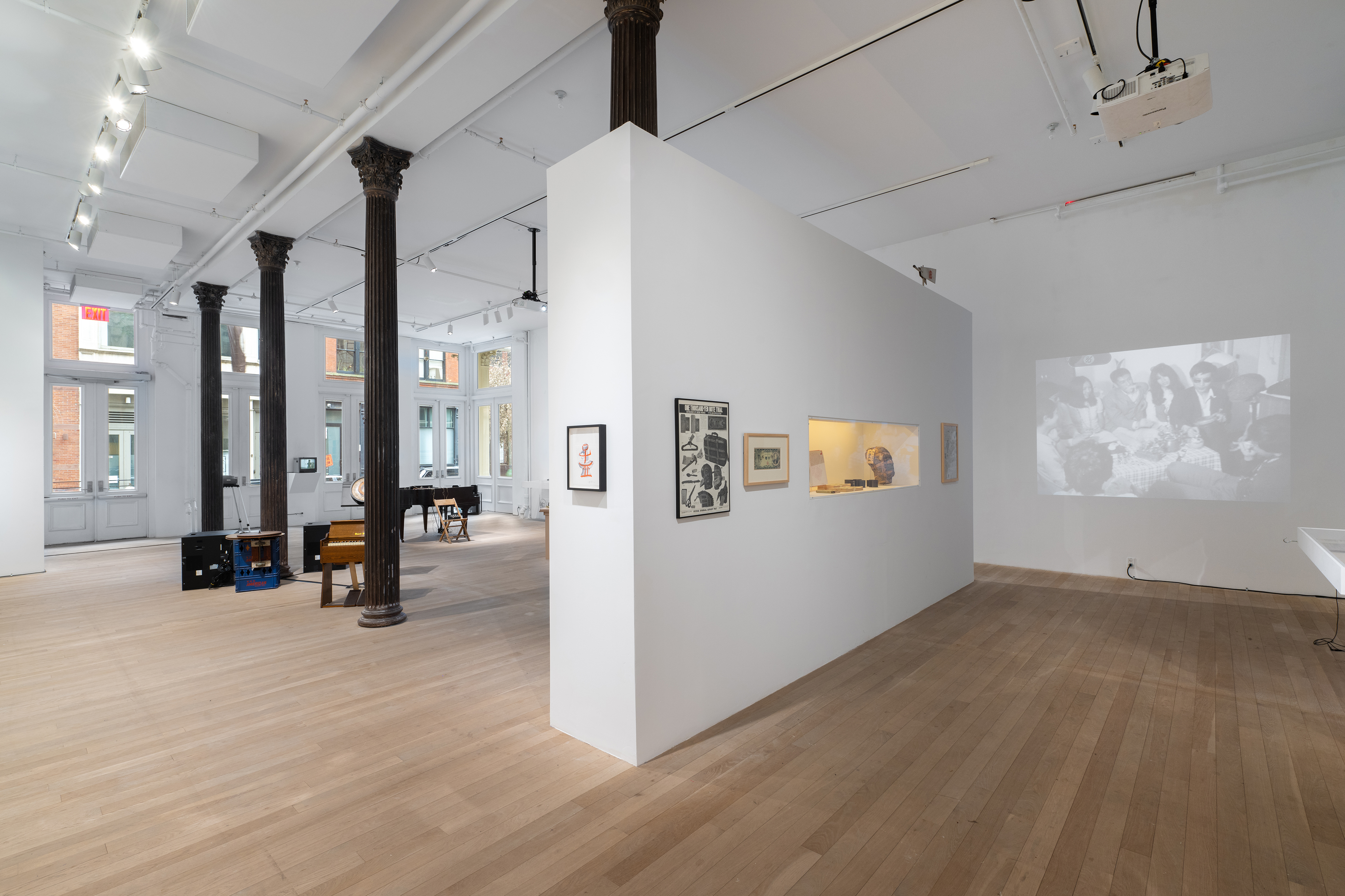 Color photograph of a gallery space. Various musical instruments and audio devices are placed around the back room, photographs and vitrines hang on the walls, and various television monitors and projections are placed along the sides of the room.