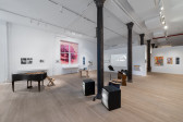 Color photograph of a gallery space. Various musical instruments and audio devices are placed around the middle of the room, photographs and vitrines hang on the walls, and various television monitors and projections are placed along the sides of the room.