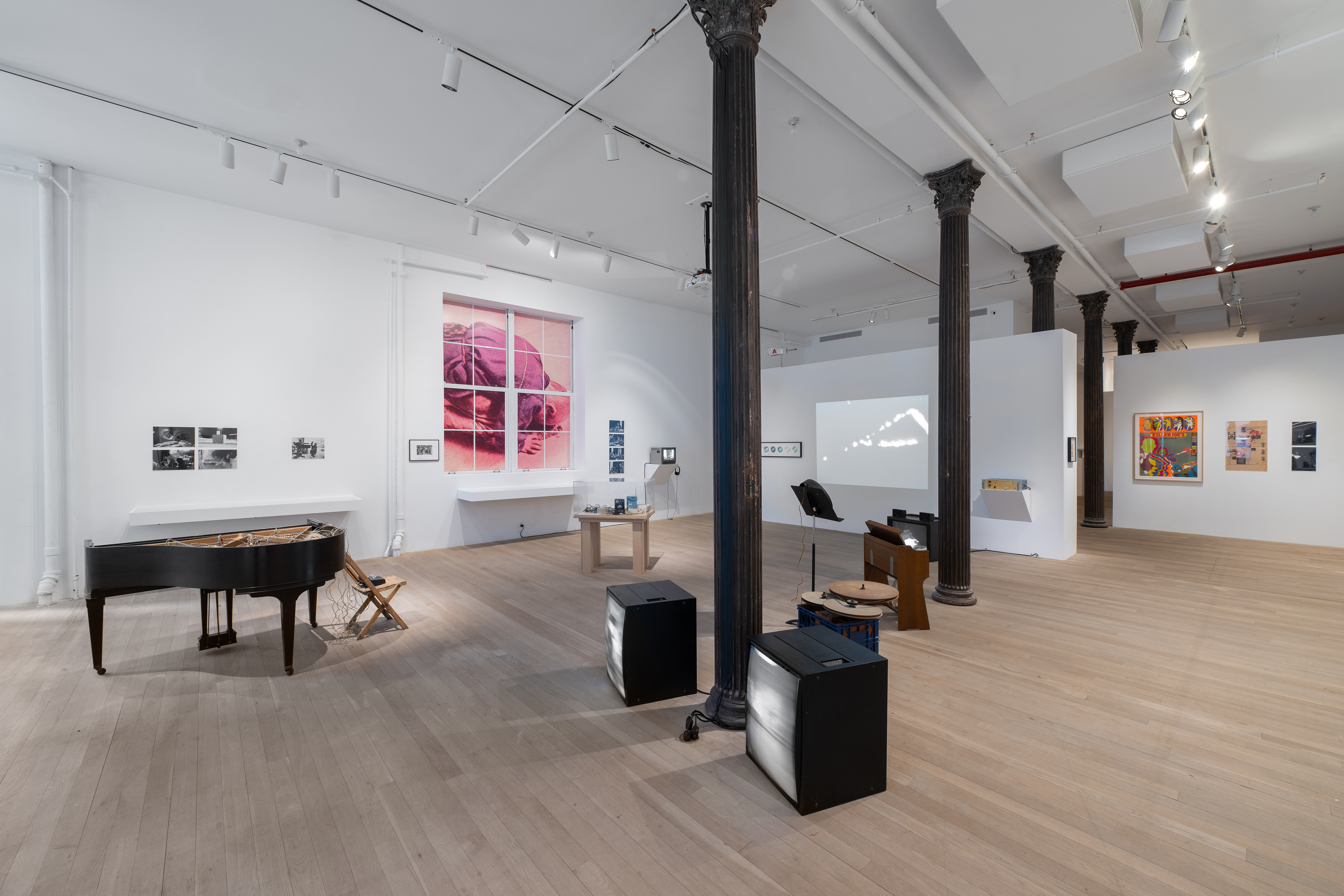 Color photograph of a gallery space. Various musical instruments and audio devices are placed around the middle of the room, photographs and vitrines hang on the walls, and various television monitors and projections are placed along the sides of the room.