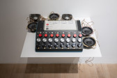 Color photograph of an audio device placed on a shelf. The device consists of a machine, speakers, and light sensors. A paper containing notes and instructions on the controls is placed on top of the machine.