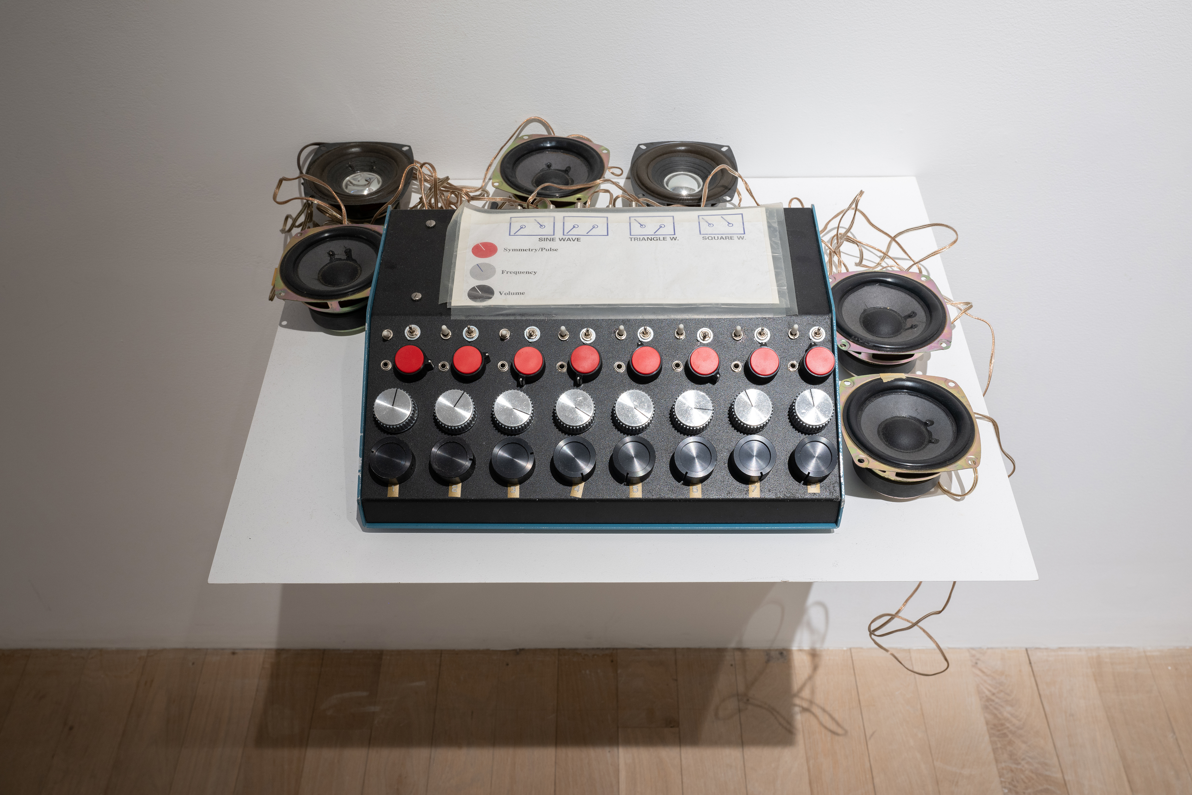 Color photograph of an audio device placed on a shelf. The device consists of a machine, speakers, and light sensors. A paper containing notes and instructions on the controls is placed on top of the machine.