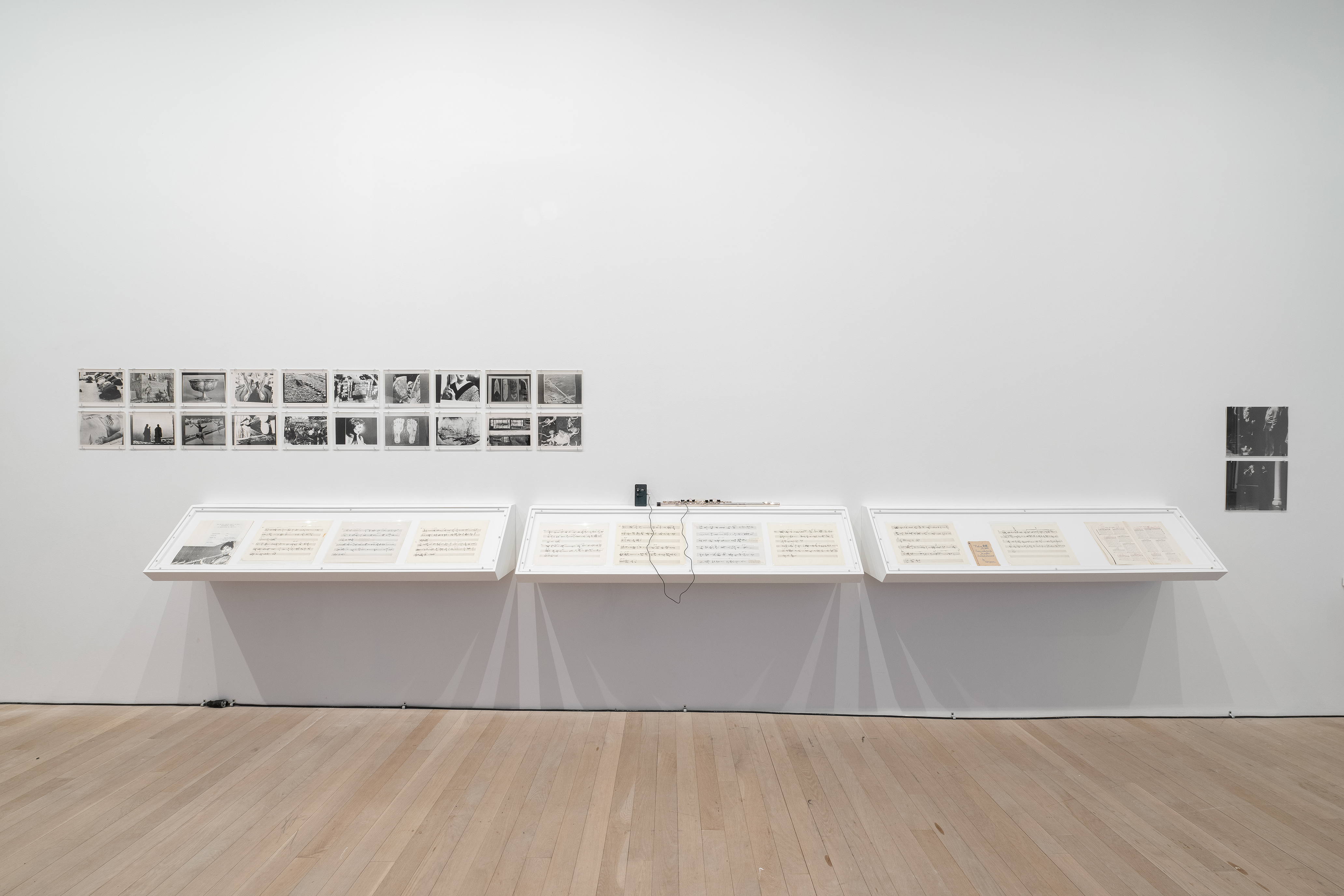 Color photograph of a white gallery wall with three vitrines containing musical scores built into the wall. On top of the vitrine is a manipulated flute, and a series of black-and-white images. Two black-and-white photographs are hung to the right of the vitrine.