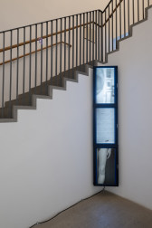 Color photograph of a gallery space. In the corner are three stacked television monitors, displaying videos taken at different speeds. On the top monitor is an image of part of a wall in a room, with a mirror and framed images. The middle monitor shows an empty image. The bottom monitor shows part of a woman's leg.