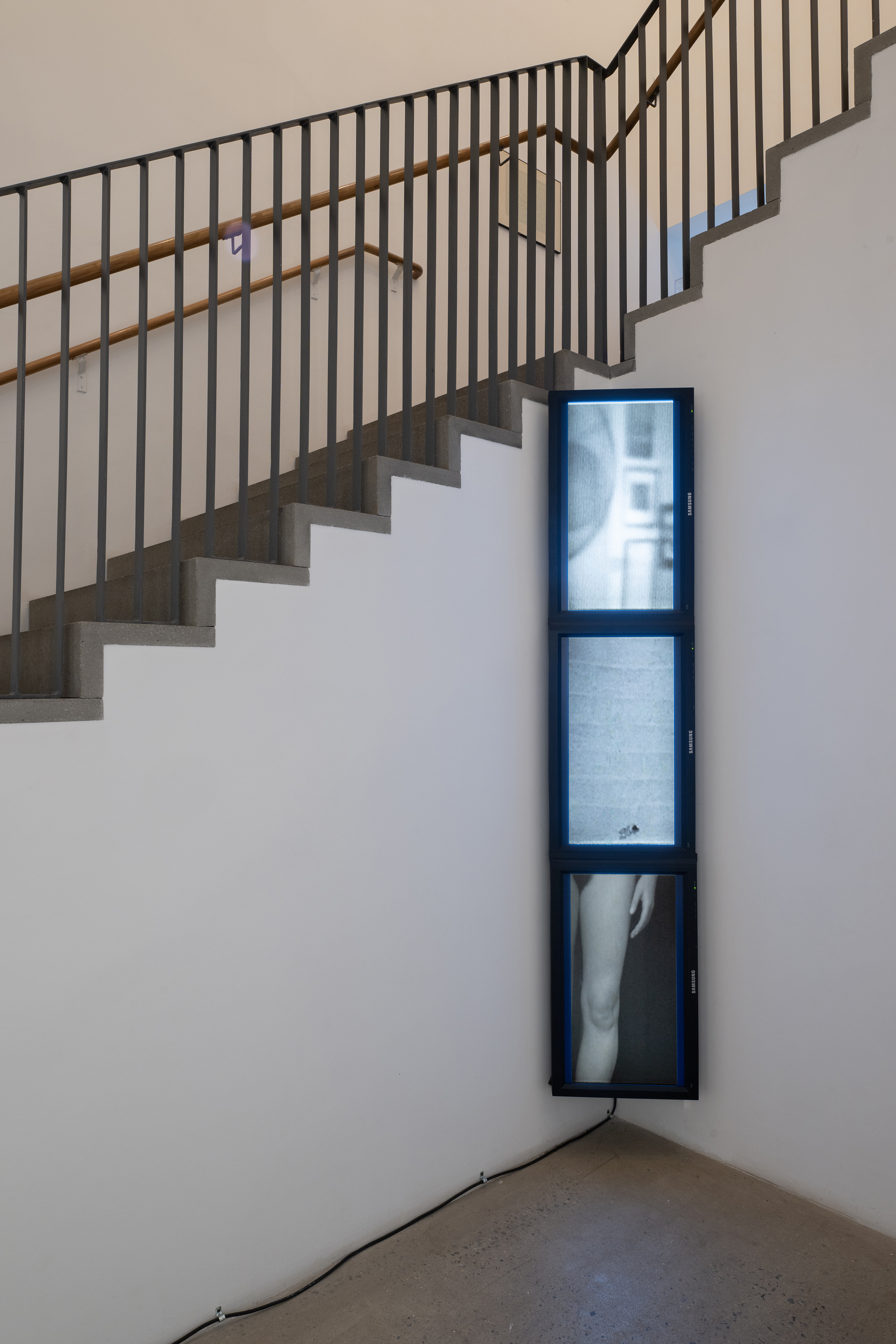 Color photograph of a gallery space. In the corner are three stacked television monitors, displaying videos taken at different speeds. On the top monitor is an image of part of a wall in a room, with a mirror and framed images. The middle monitor shows an empty image. The bottom monitor shows part of a woman's leg.