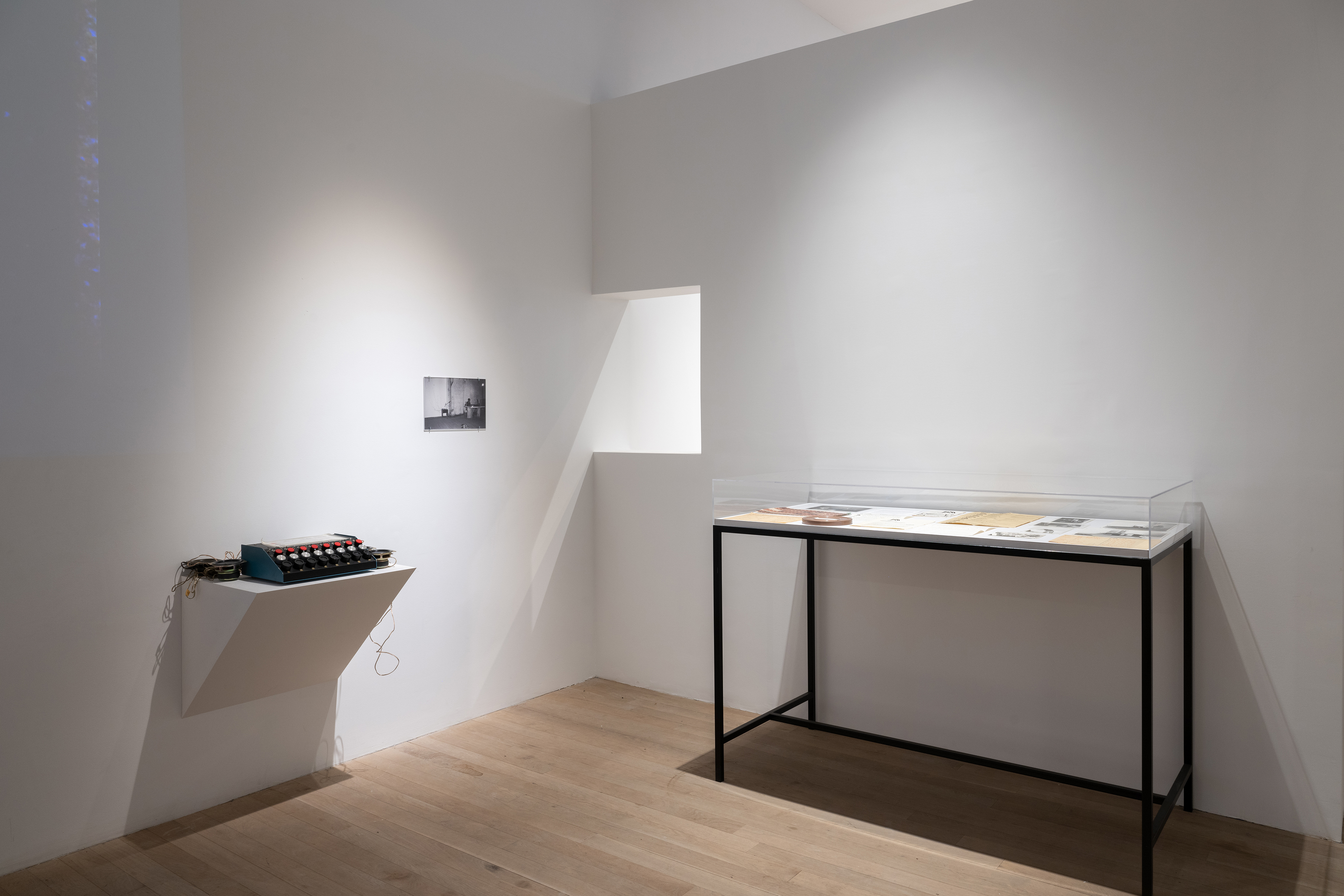 Color photograph of a corner of a gallery space. On the left wall is a shelf holding a machine, speakers, and a light sensors. A black-and-white photograph also hangs on the wall. In front of the right wall is a standing vitrine containg various ephemera.