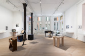 Color photograph of a gallery space. Various musical instruments and audio devices are placed around the middle of the room, photographs and vitrines hang on the walls, and various television monitors are placed along the sides of the room.