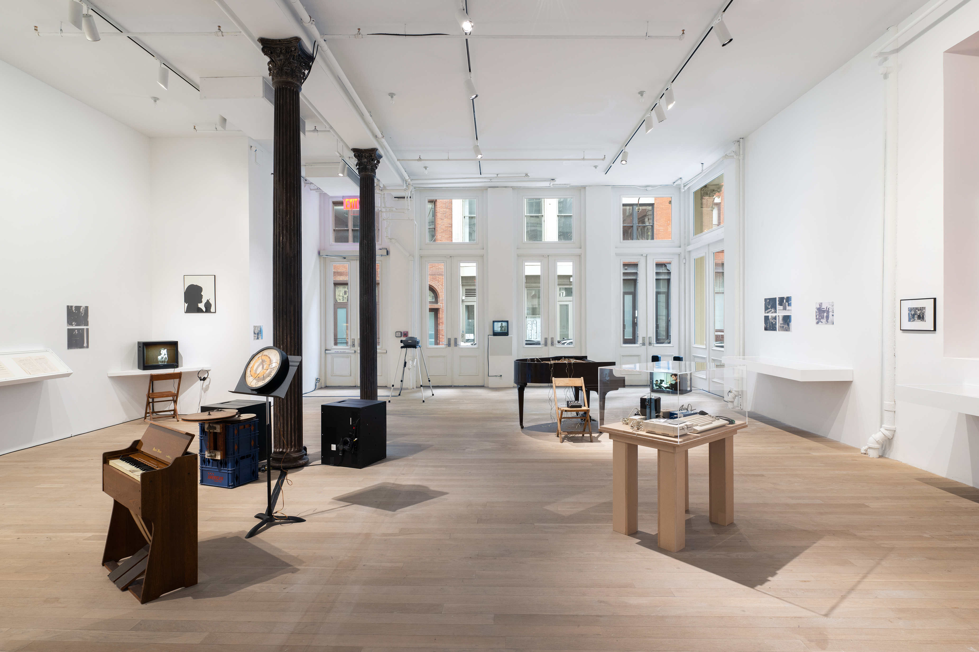 Color photograph of a gallery space. Various musical instruments and audio devices are placed around the middle of the room, photographs and vitrines hang on the walls, and various television monitors are placed along the sides of the room.