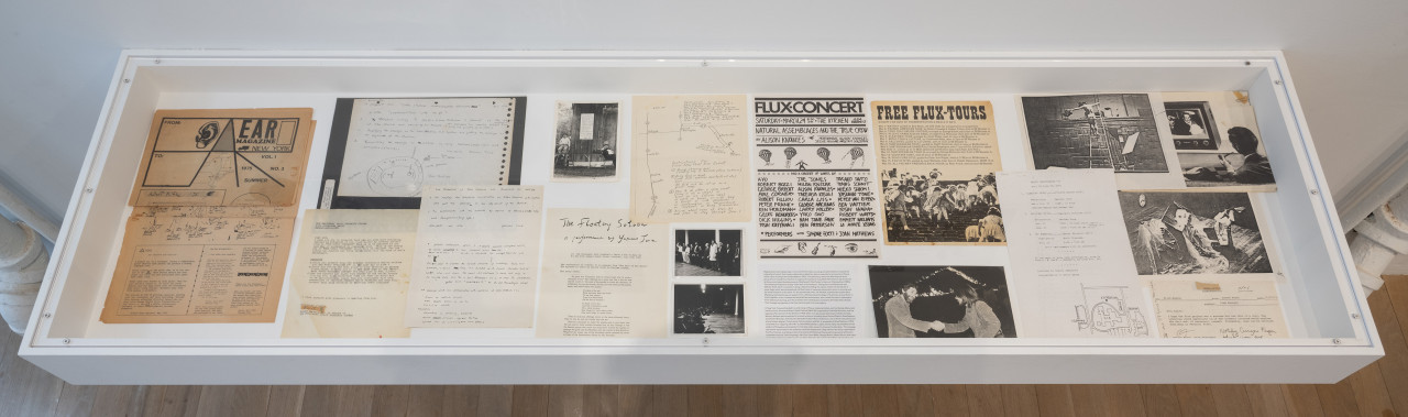 Color photograph of a vitrine from above. Various ephemera, including magazines, correspondence, and photocopied photographs in black-and-white are placed inside.