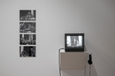 Color photograph of a gallery wall. On the left, four black-and-white photographs are hung vertically, and on the right is a shelf with a television monitor placed on it.