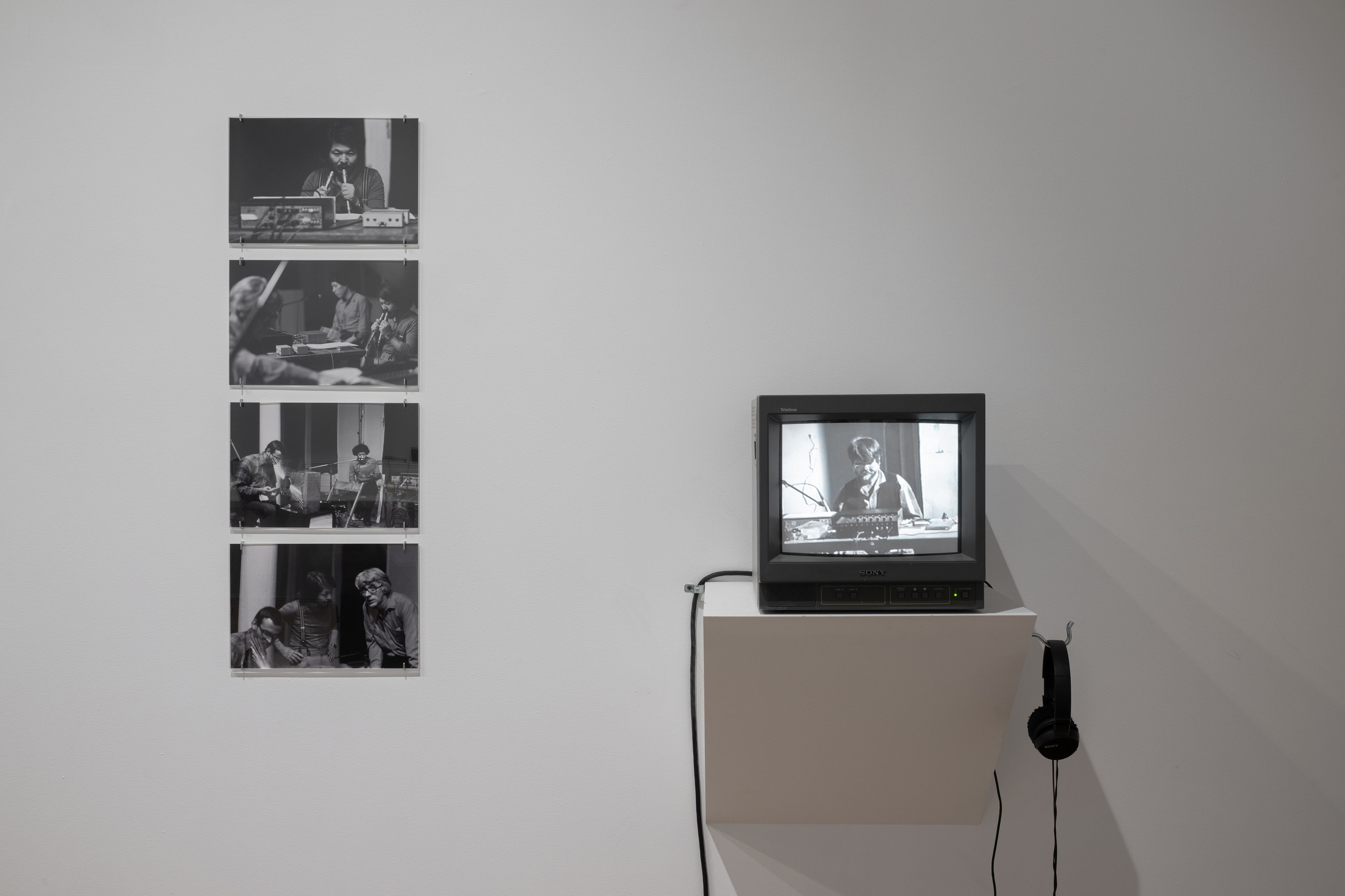 Color photograph of a gallery wall. On the left, four black-and-white photographs are hung vertically, and on the right is a shelf with a television monitor placed on it.