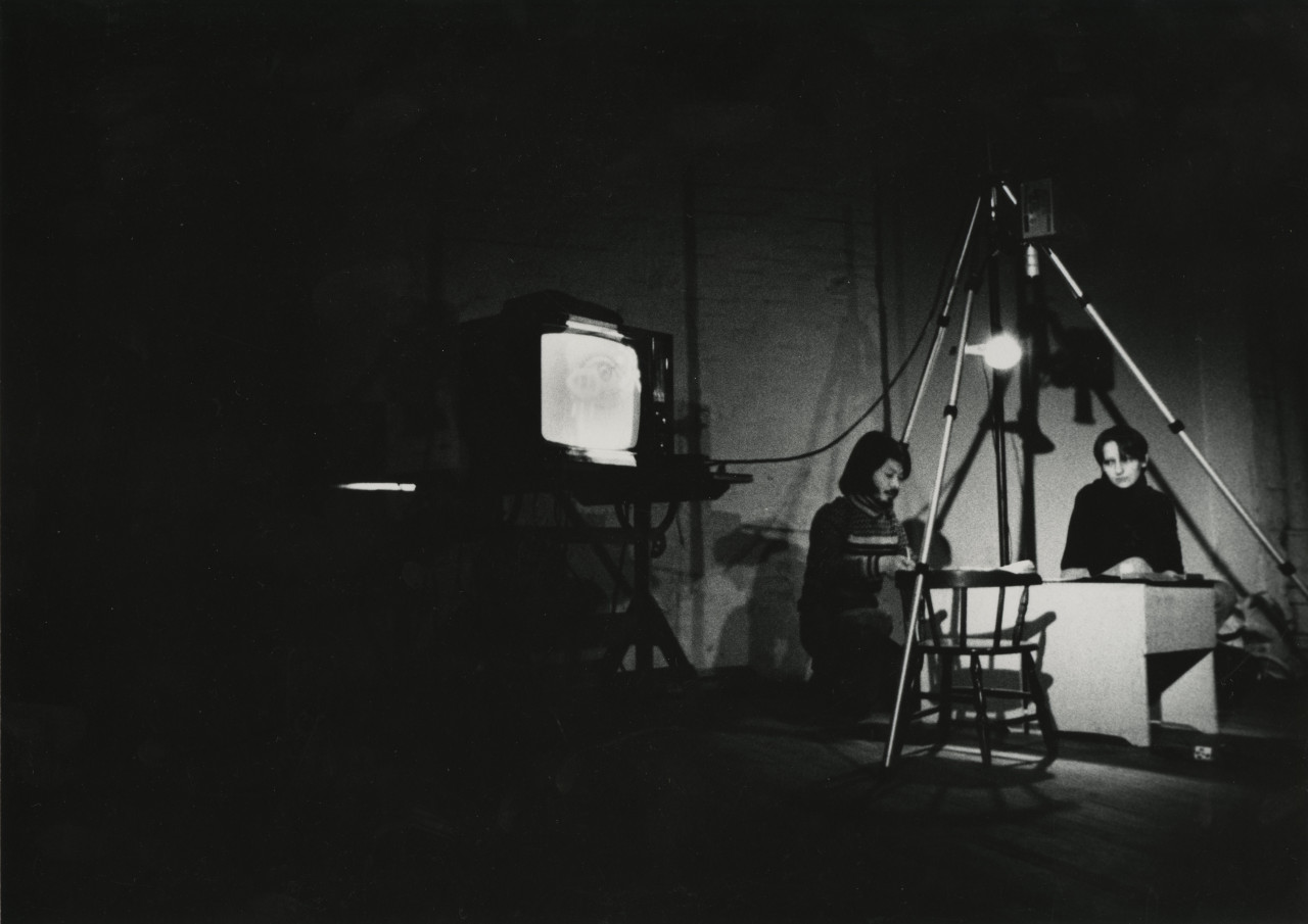 Black-and-white photograph of a TV against a brick wall, connected to a camera recording directly above a spotlighted chair and desk. A figure sits across the empty chair at the desk, while another figure kneels down to the left side of the desk working on something.