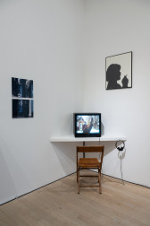 Color photograph of a corner of a gallery space. On the left wall are two black-and-white photographs, and on the right wall is a black-and-white silhouette of a figure smoking. In the corner, a television monitor sits on a shelf.
