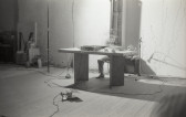 Black-and-white photograph of a blurred figure in motion sitting behind a wooden desk surrounded by audio equipment in a sparse room.