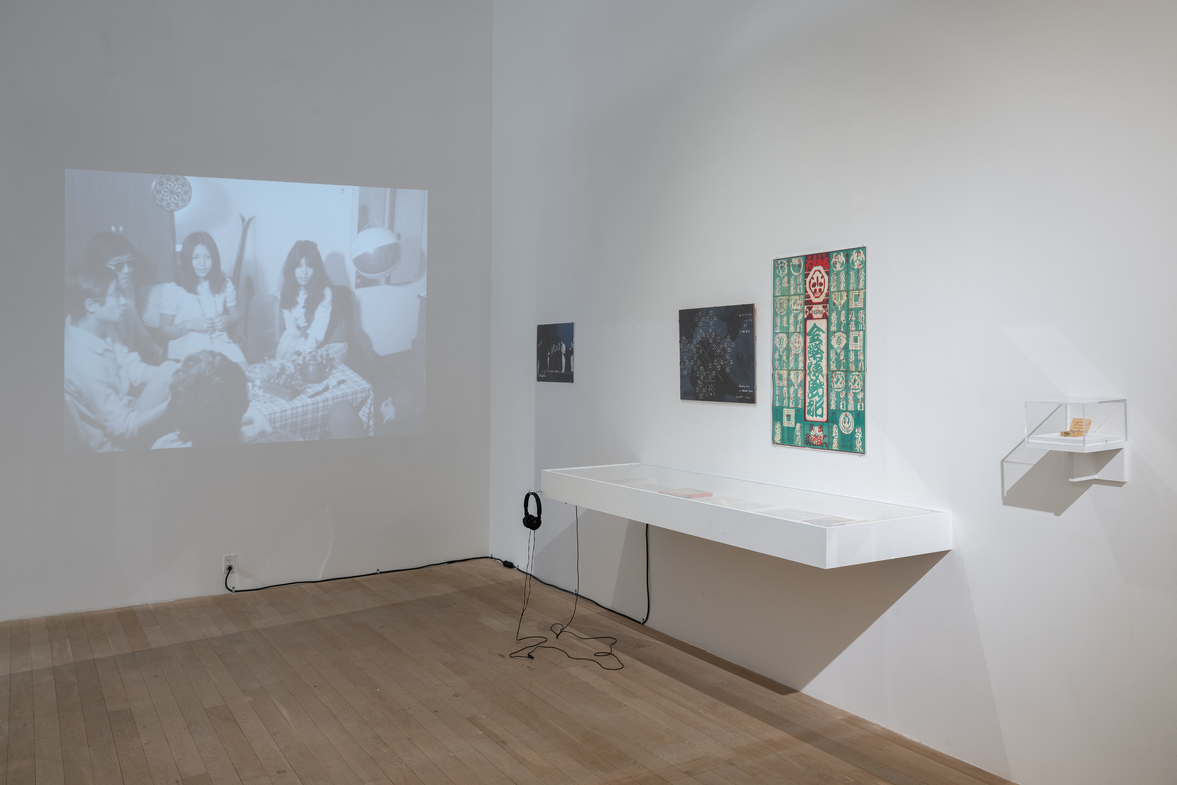 Color photograph of a gallery space. On the left wall is a projected black-and-white video. On the right wall are various scores, posters, and photographs, and a vitrine.