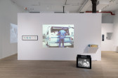 Color photograph of a gallery space. A free-standing wall sits in the middle of the space, holding framed CDs, a projected image of a man and a truck, and an audio device. On the floor in front of the wall sits a television monitor.
