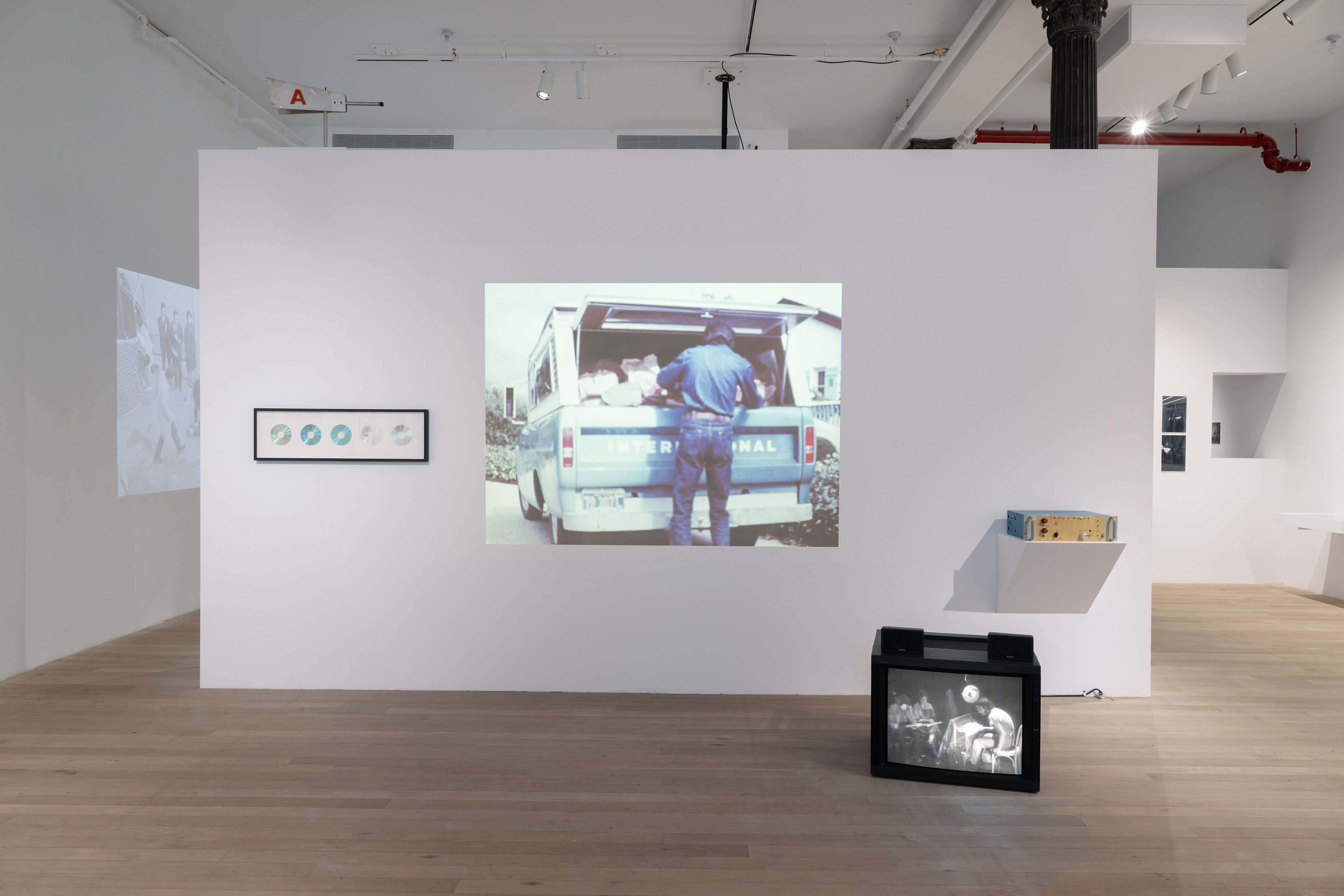 Color photograph of a gallery space. A free-standing wall sits in the middle of the space, holding framed CDs, a projected image of a man and a truck, and an audio device. On the floor in front of the wall sits a television monitor.