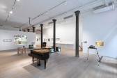 Color photograph of a gallery space. Various musical instruments and audio devices are placed around the middle of the room, photographs and vitrines hang on the walls, and various television monitors and projected images are placed along the sides of the room.