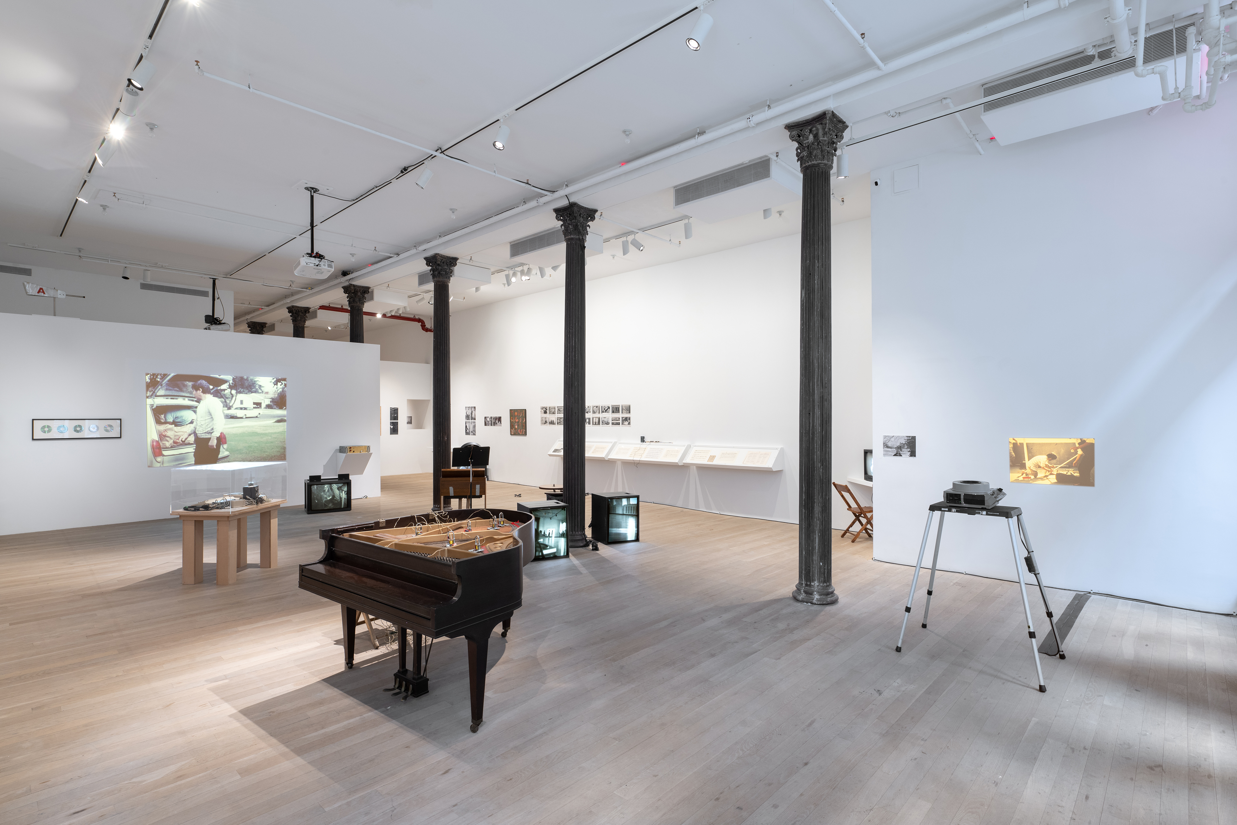 Color photograph of a gallery space. Various musical instruments and audio devices are placed around the middle of the room, photographs and vitrines hang on the walls, and various television monitors and projected images are placed along the sides of the room.