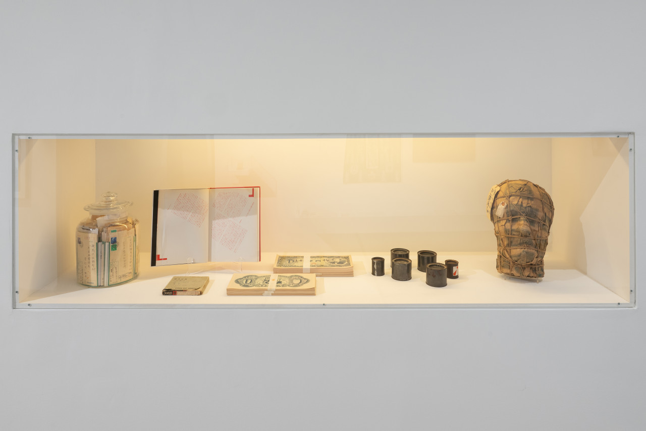 Color photograph of a vitrine set into a wall. Placed inside are various works including a book, stacks of printed imitation yen sheets, film cannisters, and a mask made of printed imitation yen sheets.