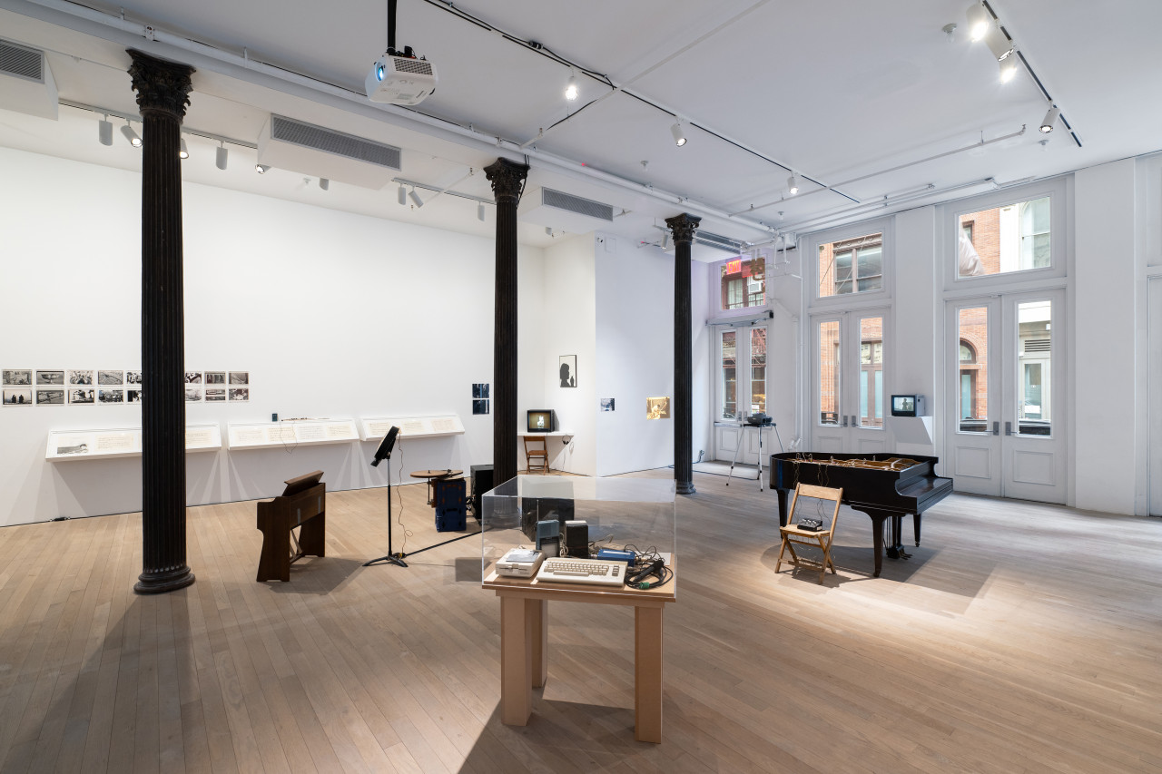 Color photograph of a gallery space. On the left wall series of photographs hang, and vitrines containing musical scores. Various musical instruments and audio devices are placed around the middle of the room, and various television monitors are placed along the sides of the room..